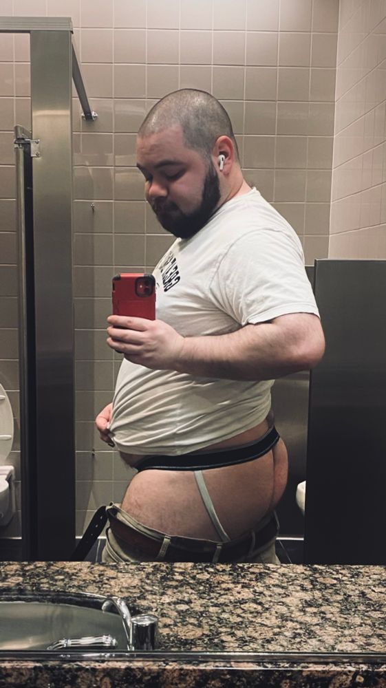 A selfie of me in a public restroom. My pants are unbuckled and lowered and I’m turned at an angle, showing off my ass and jockstrap. band of the strap dips be