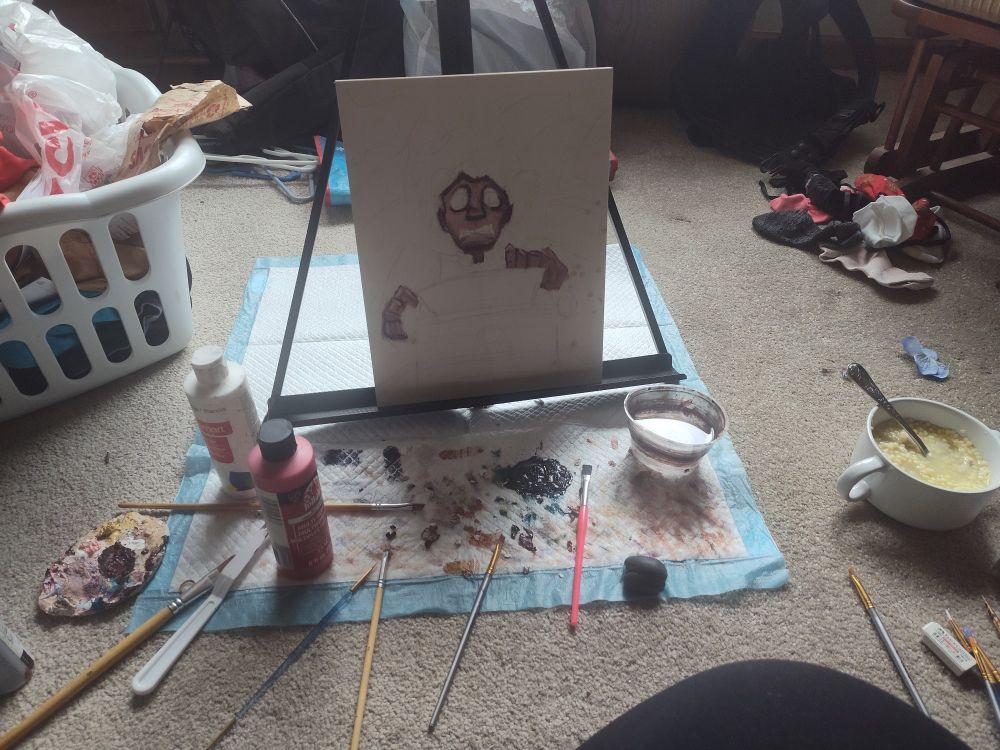 A messy room. In the center is an easel with a barely painted wooden board on it. Surrounding it is a few paint brushes, a cup of water, paint, a kneadable eraser, and a bowl of chicken noodle soup.