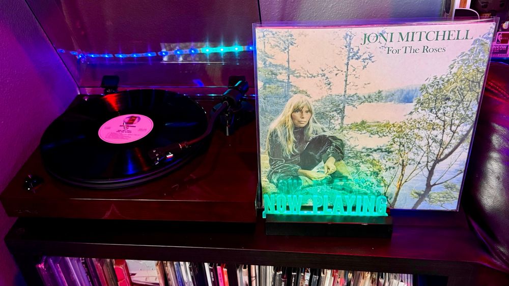 A Fluance RT82 turntable plays a vinyl record with a white Reprise label. To the right, a glowing cyan "NOW PLAYING" stand displays the album "For the Roses" by Joni Mitchell. The cover art shows Joni seated in a forested area beside a lake, with trees in the background. She rests her hands on her knees and gazes calmly ahead, wearing a dark outfit and moccasin-style boots. The scene is tranquil and introspective, matching the tone of the music.