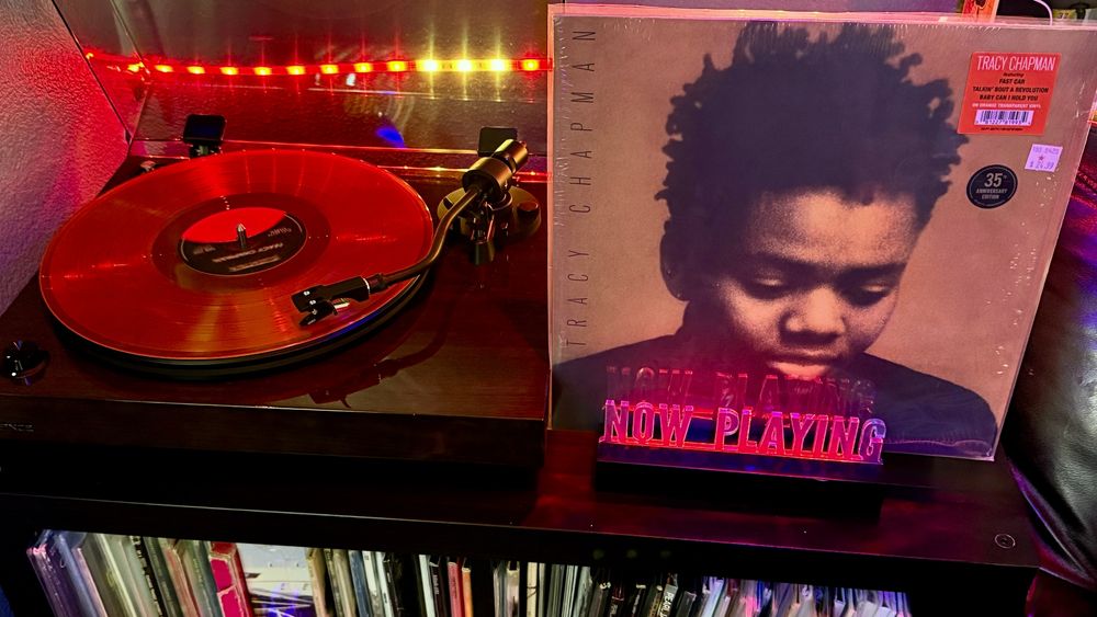 A vibrant, orange transparent vinyl record is spinning on a sleek, dark wood turntable with a clear dust cover lifted open. The tonearm is engaged, playing the record. To the right, the album cover for Tracy Chapman's self-titled debut album is prominently displayed in a protective sleeve. The cover features a sepia-toned close-up portrait of Tracy Chapman with eyes cast downward and natural, textured hair. A red sticker on the plastic wrap highlights the 35th Anniversary Edition and mentions hit tracks like “Fast Car,” “Talkin’ Bout a Revolution,” and “Baby Can I Hold You.” A red LED acrylic sign in front of the album reads “NOW PLAYING.” The background is bathed in warm red and purple LED lighting, and beneath the turntable is a shelf holding more vinyl records in upright sleeves, showcasing a curated collection.