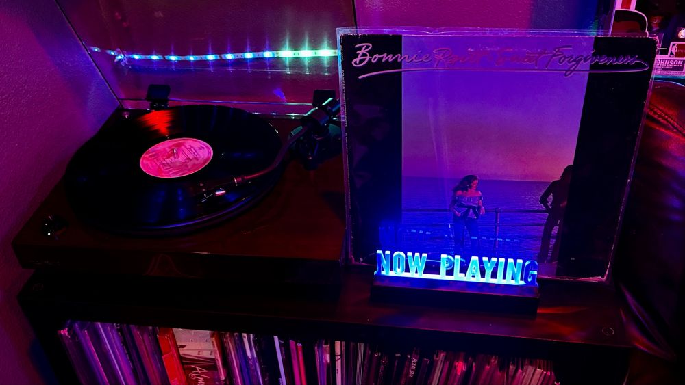 A black vinyl record spins on a Fluance RT82 turntable with a glossy dark wood finish, under ambient magenta and blue LED lighting. To the right of the turntable, the album cover for Bonnie Raitt – Sweet Forgiveness is displayed upright in a plastic sleeve. The cover shows Bonnie Raitt standing on a boardwalk near the ocean during sunset, with another figure leaning on the railing in silhouette. Above them, her name and the album title are written in a flowing script. In front of the album is a glowing blue acrylic sign that reads “NOW PLAYING.” Below the turntable setup is a shelf filled with vinyl records, neatly stored upright.