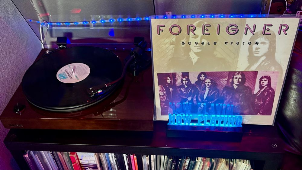 A black vinyl record spins on a dark wood turntable with a clear acrylic dust cover propped open. To the right of the turntable, the album cover for Foreigner – Double Vision is displayed upright in a plastic sleeve. The cover features a large faded photo of two band members in the background and a smaller, more prominent band photo in a reddish tone along the lower half, showing the full group in leather jackets. In front of the album, a glowing blue acrylic sign reads “NOW PLAYING.” The background is lit with blue LED strip lights, and below the setup is a shelf with neatly stored vinyl records in upright sleeves, revealing a portion of a larger music collection.