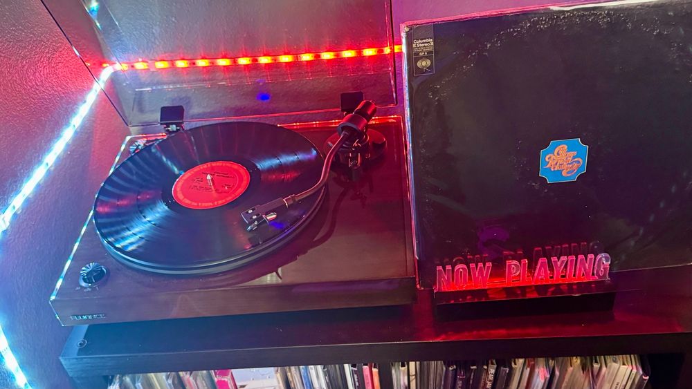 A Fluance RT82 turntable spins a record with a red Columbia label, surrounded by vibrant red and blue LED lighting. To the right, a red-glowing "NOW PLAYING" stand holds the Chicago Transit Authority album by Chicago. The iconic debut album cover is stark and minimalistic—solid black with a small ornate blue and gold logo reading "Chicago Transit Authority" centered toward the top. The plastic sleeve shows signs of age and wear, adding a vintage charm to the setup. Vinyl records are neatly shelved below.