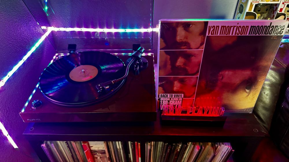 A Fluance RT82 turntable spins a vinyl record with a light brown Warner Bros. label under vibrant, multicolored LED lights casting purples, blues, and greens across the setup. To the right, a glowing red “NOW PLAYING” sign supports the Moondance album by Van Morrison, featuring a collage of the artist’s face in warm amber tones. A sticker on the album sleeve reads “BACK TO VINYL - ORIGINAL ALBUM ON 180-GRAM VINYL.” The turntable and display rest atop a media console filled with more vinyl records, with a few Funko Pop! figures visible on the shelf above. The cozy lighting and thoughtful arrangement create a nostalgic, audiophile vibe.