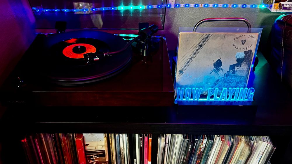A Fluance RT82 turntable in a dark wood finish plays a 7-inch vinyl record with a bright orange and black label under vivid blue and purple LED lighting. To the right of the turntable, the album cover for Pearl Jam’s Merkinball EP is displayed upright. The minimalist cover features a crane lifting a satellite dish above a building, with the band’s name and EP title handwritten in black marker along with a small black heart. A vintage-looking price sticker reads “$3.00.” A glowing blue acrylic “NOW PLAYING” sign sits in front of the record. The shelf below is filled with neatly organized vinyl albums.