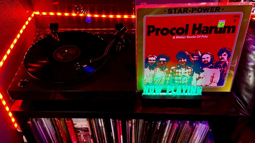 A Fluance RT82 turntable is playing a vinyl record under vibrant red LED lighting that lines the corner of the setup. Next to the turntable is a glowing 'NOW PLAYING' sign illuminated in green, placed in front of the album cover for Procol Harum's A Whiter Shade of Pale, part of the 'Star-Power' series. The album features a stylized illustration of the band on a bold red and gold background. Below the setup is a shelf filled with neatly organized vinyl records.