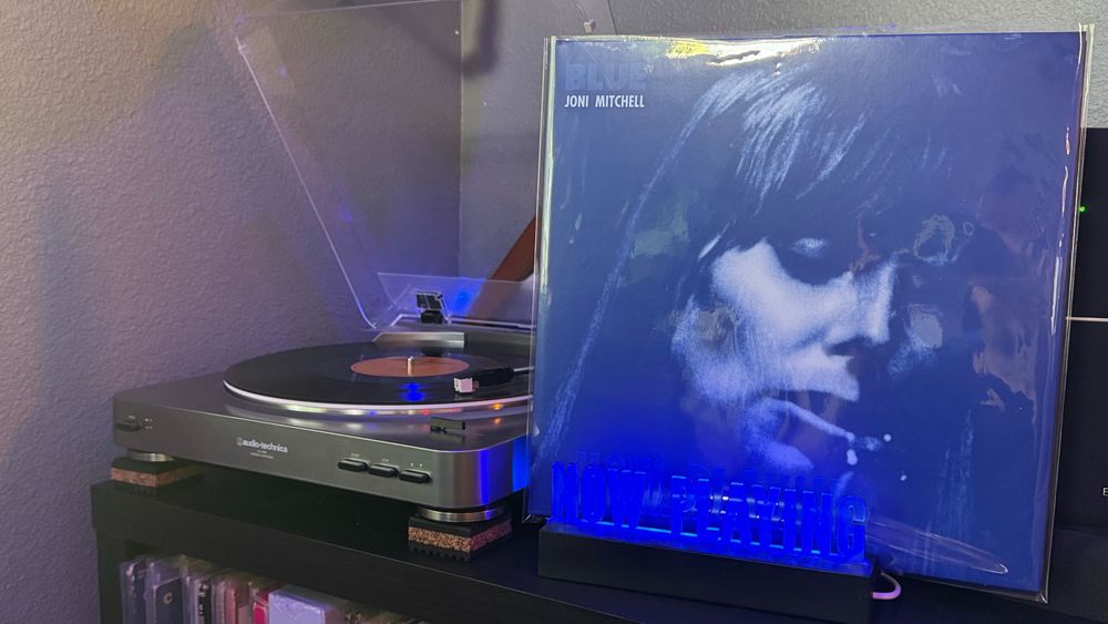 A vinyl record of "Blue" by Joni Mitchell spins on an Audio-Technica turntable, with the album cover displayed upright in a blue-lit “Now Playing” stand. Below the setup is a shelf filled with vinyl records, and the scene is warmly lit, evoking a cozy listening environment.