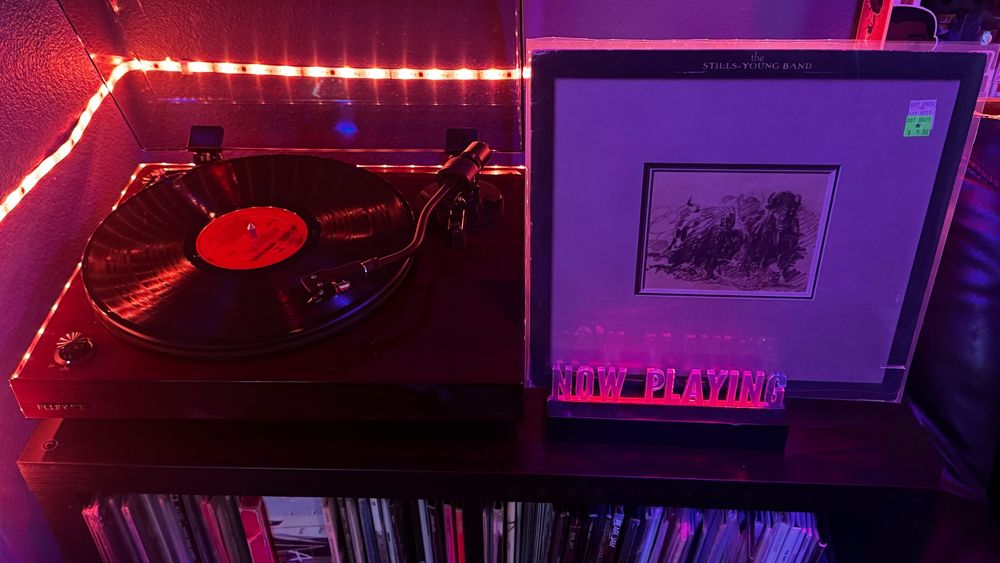 A Fluance RT82 turntable plays a vinyl record with a red label under warm red LED lighting. To the right, a red-lit "NOW PLAYING" stand displays the album Long May You Run by The Stills-Young Band. The minimalist cover features a grayscale sketch of two wolves or huskies pulling a sled in rough terrain, framed against a stark white background. A small green price sticker in the upper right corner shows it was purchased for $5.00. The setup exudes a vintage, analog vibe with a cozy, dimly lit ambiance.