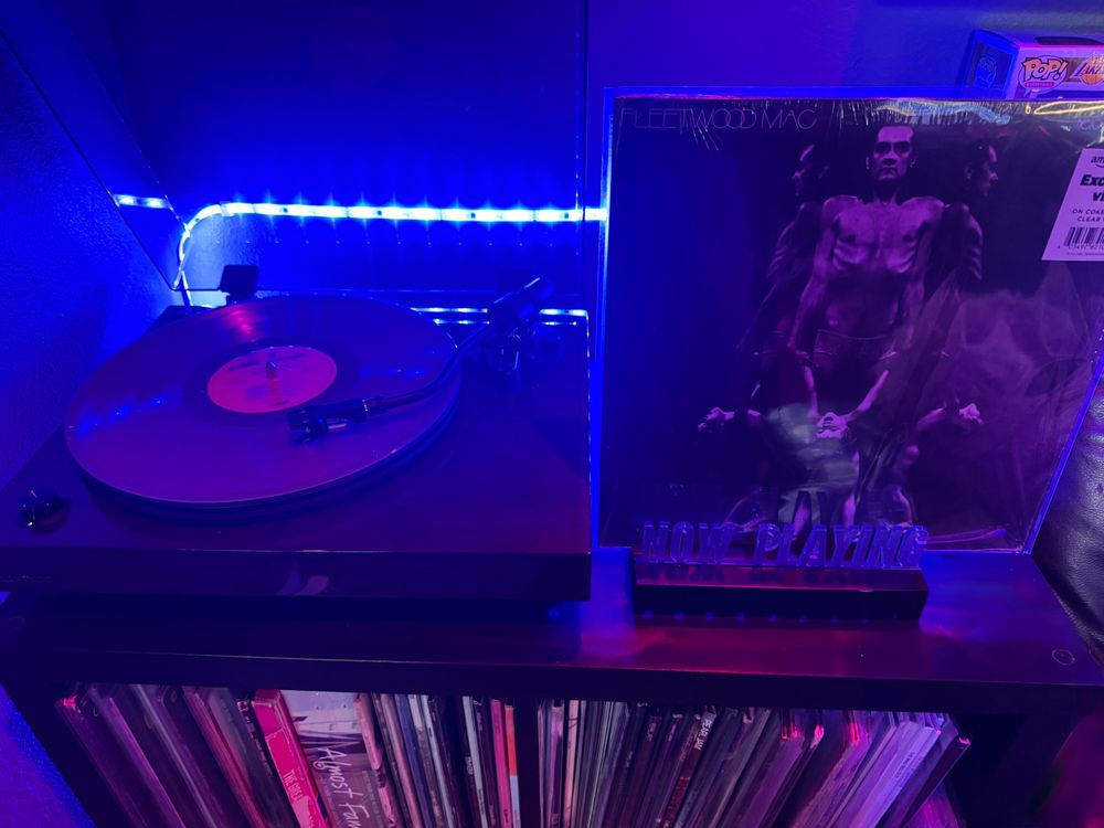  A Fluance RT82 turntable spinning Fleetwood Mac’s Heroes Are Hard to Find on coke‑bottle clear vinyl. Beside it, the album cover stands in a “Now Playing” display: a three‑way mirrored black‑and‑white photo of Mick Fleetwood—chest puffed out and ribs showing—standing in lace underpants, holding his young daughter Amelia’s hands as she stands on his shoes. The moody scene is bathed in blue and purple LED lighting, with a vinyl-filled shelf below.