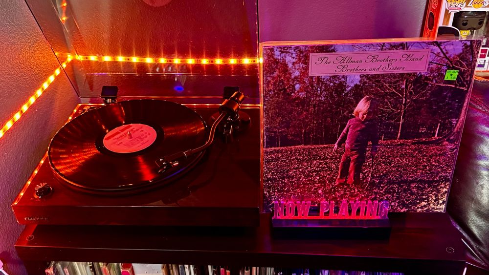 A Fluance RT82 turntable spins a vinyl record with a white Capricorn Records label under warm orange LED lighting. Next to it, an illuminated “NOW PLAYING” sign glows red beneath the album cover for Brothers and Sisters by The Allman Brothers Band, which shows a young blond child standing alone in a field of autumn leaves. The setup rests atop a black media shelf filled with neatly arranged vinyl records. A few Funko Pop figures are visible in the top-right corner, adding a personal touch to the listening space.