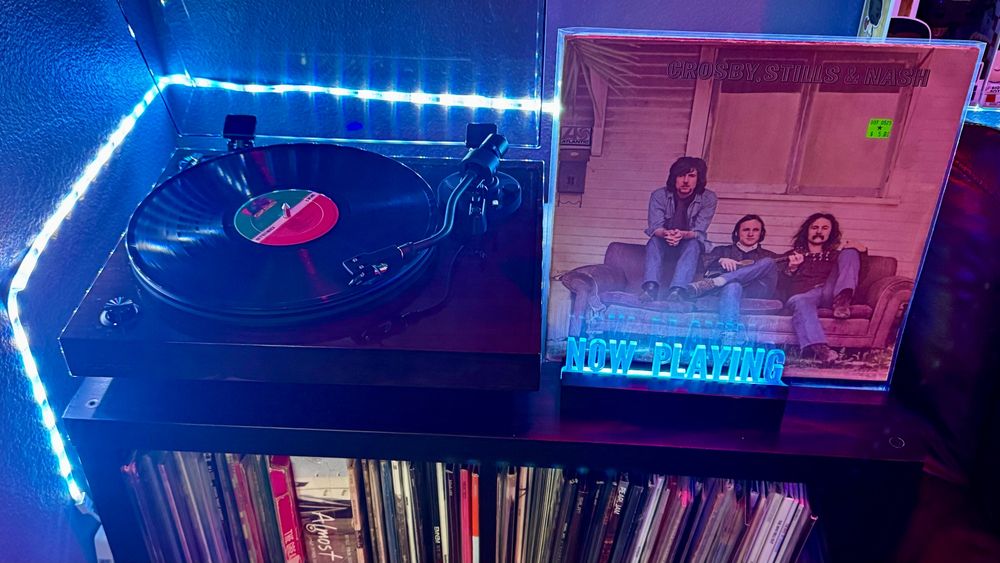 A Fluance RT82 turntable is spinning a vinyl record under cool blue LED lighting that outlines the corner of the setup. Next to the turntable is a bright blue 'NOW PLAYING' sign, placed in front of the album cover for Crosby, Stills & Nash. The cover features the three band members sitting casually on a worn-out couch in front of a weathered wooden house. Below the setup, shelves are filled with a neatly arranged collection of vinyl records, with a visible spine for the Almost Famous soundtrack among them.