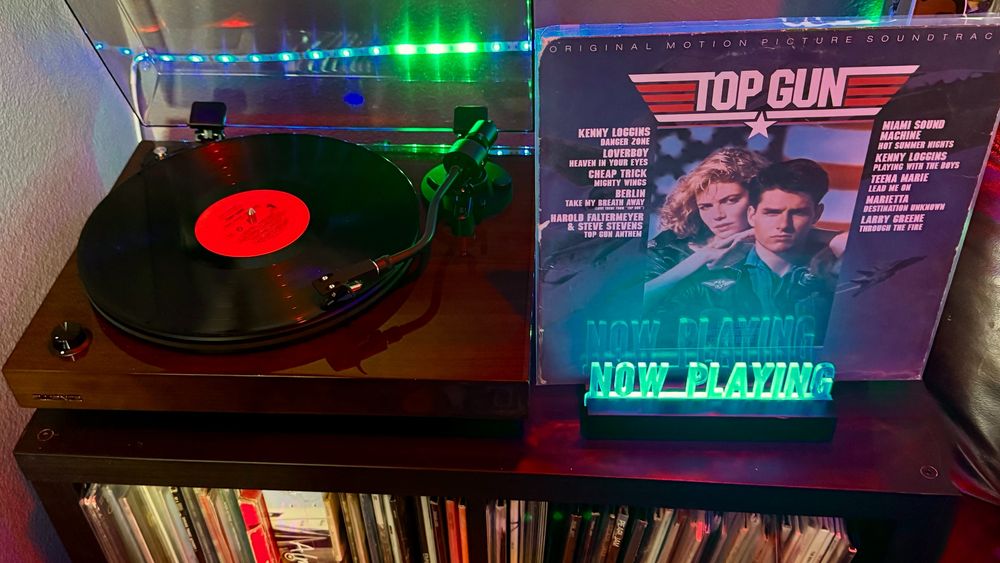 A Fluance turntable is playing a vinyl record with a bright red center label, illuminated by cool green and blue LED lights mounted along the back wall. To the right of the player, the Top Gun Original Motion Picture Soundtrack album is proudly displayed in its sleeve. The iconic cover features actors Tom Cruise and Kelly McGillis in a dramatic pose with a fighter jet soaring in the background. The "Top Gun" title is styled in bold white and red with a star emblem beneath it. Listed on the cover are artists featured on the soundtrack, including Kenny Loggins, Berlin, Cheap Trick, and more. In front of the album sits a glowing green "NOW PLAYING" acrylic sign. Vinyl records are neatly organized on the shelf below the setup.