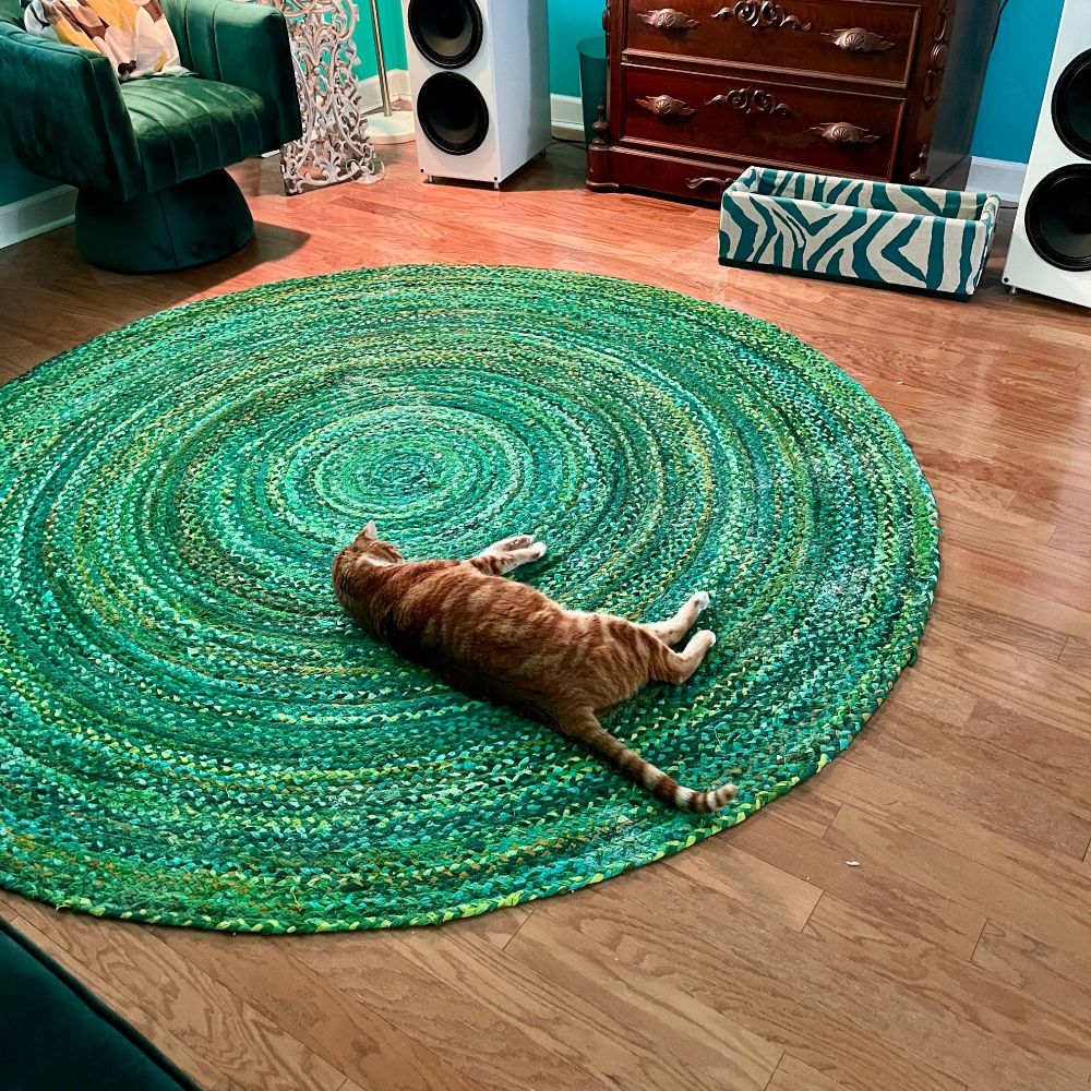 Stripey orange cat sprawled on a bright multi-green braided cotton rug. We do not know why. Lobster, what r u doing.