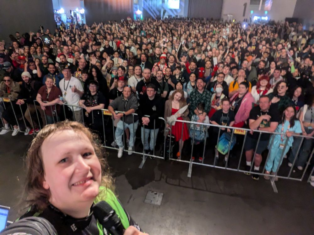 On stage at PAX Australia, Triana takes a selfie with the entire expo queue hall in the background. Triana is wearing a black and green Ground Zero Esports jersey and holding a microphone.