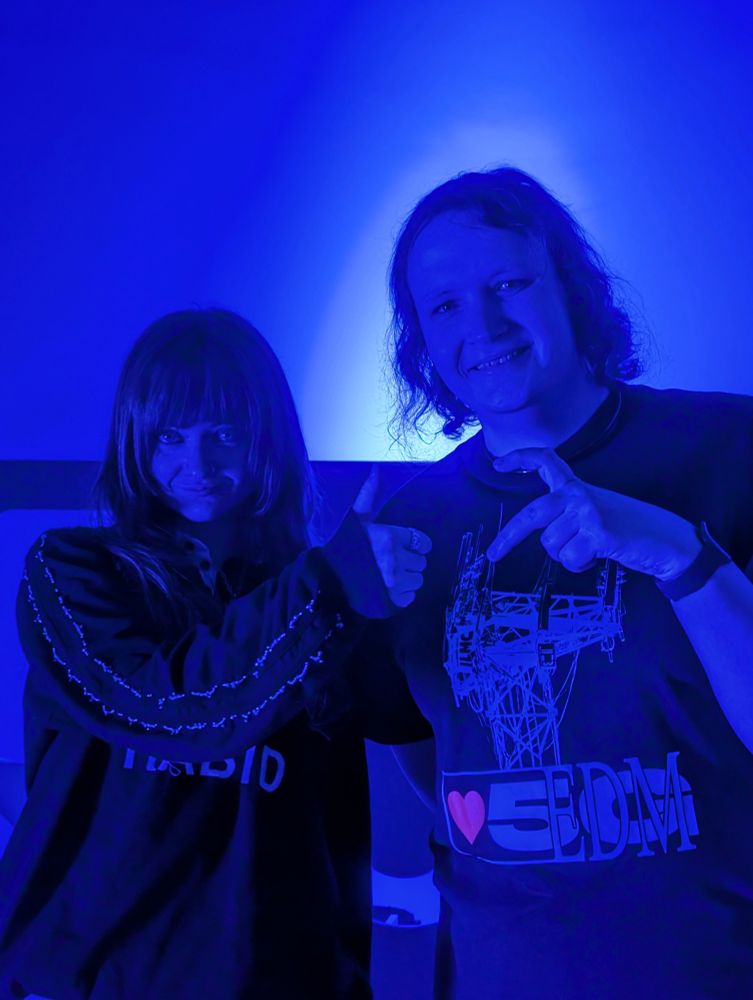Nina and Triana standing in royal blue light in a studio. Nina wears a sports jersey which says "rabid" across it. Triana wears a Ninajirachi I Heart 5G shirt with a cell tower on it. Triana is doing one half of a heart symbol with its hands, and instead of doing the other half, Nina is doing a thumbs up.