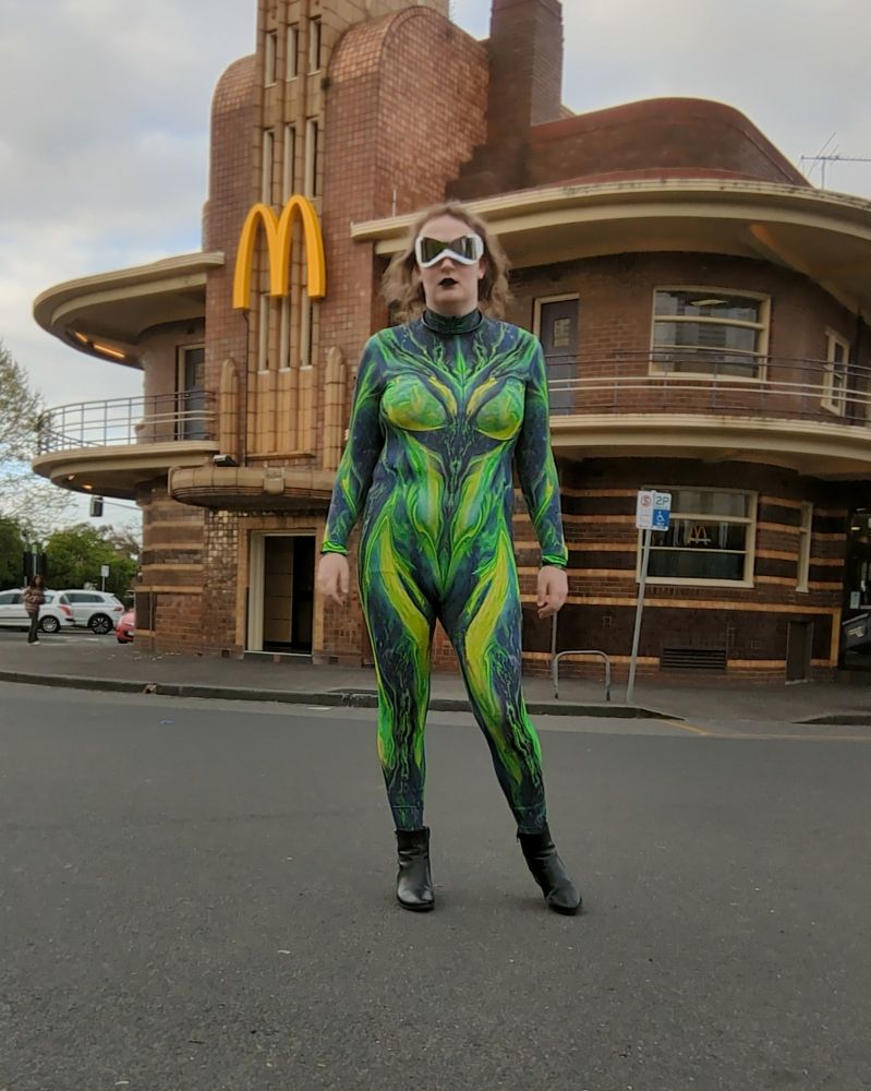 Triana wearing a toxic green coloured full body zentai suit, with energetic patterns flowing around its chest and thighs, paired with black lipstick and silver face-wrapping sunglasses. In the background, the art deco McDonald's in Clifton Hill makes the whole photo look like it was shot in an alternate universe. zentai bodysuit by Badinka.