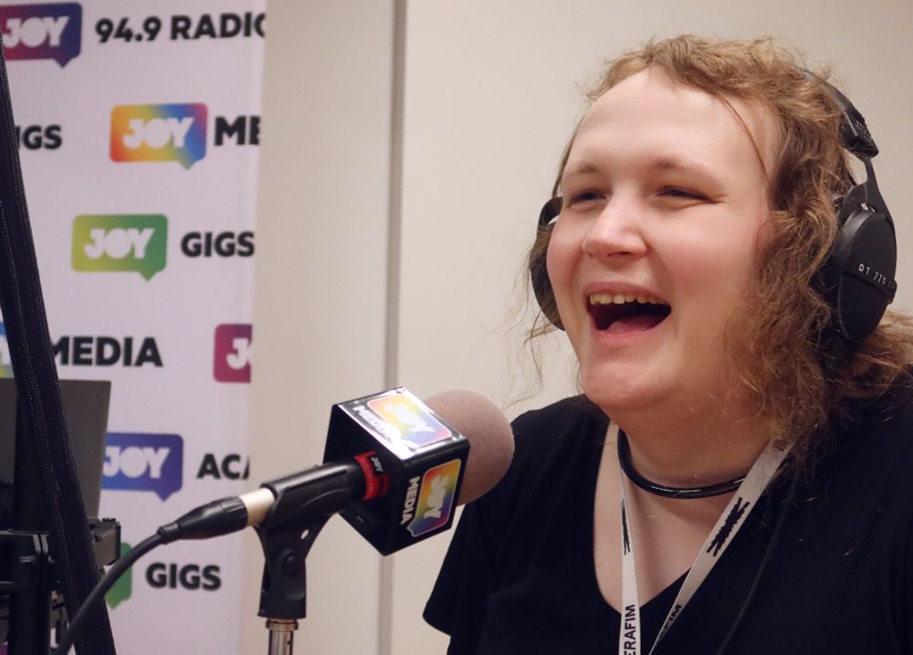 In a radio studio, Triana laughs hard on mic. She's wearing some Beyer Dynamic headphones, a Le Sserafim lanyard, and her black collar that matches her black dress.