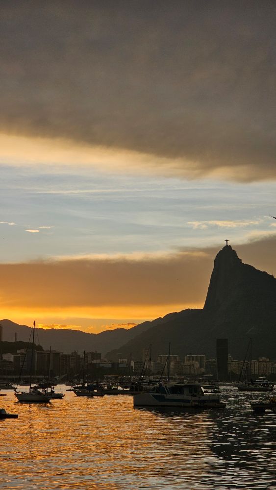 O cristo redentor, o corcovado, o céu azul e laranja, a baía de guababara com uns barcos e os prédios