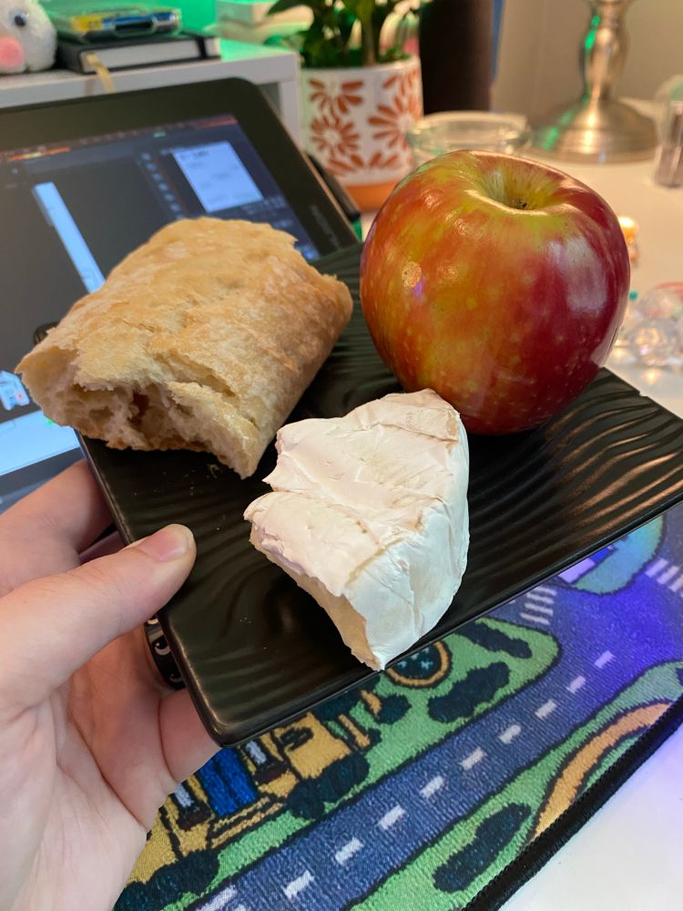 a photo of a black square plate, matte finish and wood grain texture, holding a roughly torn section of baguette, a hastily sectioned wedge of brie, and a whole apple. a pale hand holds the place over a white desk, and a desk mat made to look like those classic rugs for kids with roads and towns on them. a drawing tablet and various desk trinkets can be seen in the background.
