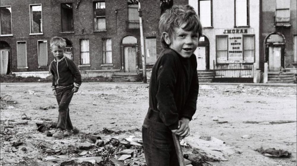 A photo taken from “Dublin, The Heart of the City”, by Ronan Sheehan and Brendan Walsh. A pre-apocalyptic scene from the 1980s: two cheery boys stand on waste land in front of a row bricked-up/collapsed Georgian houses. 