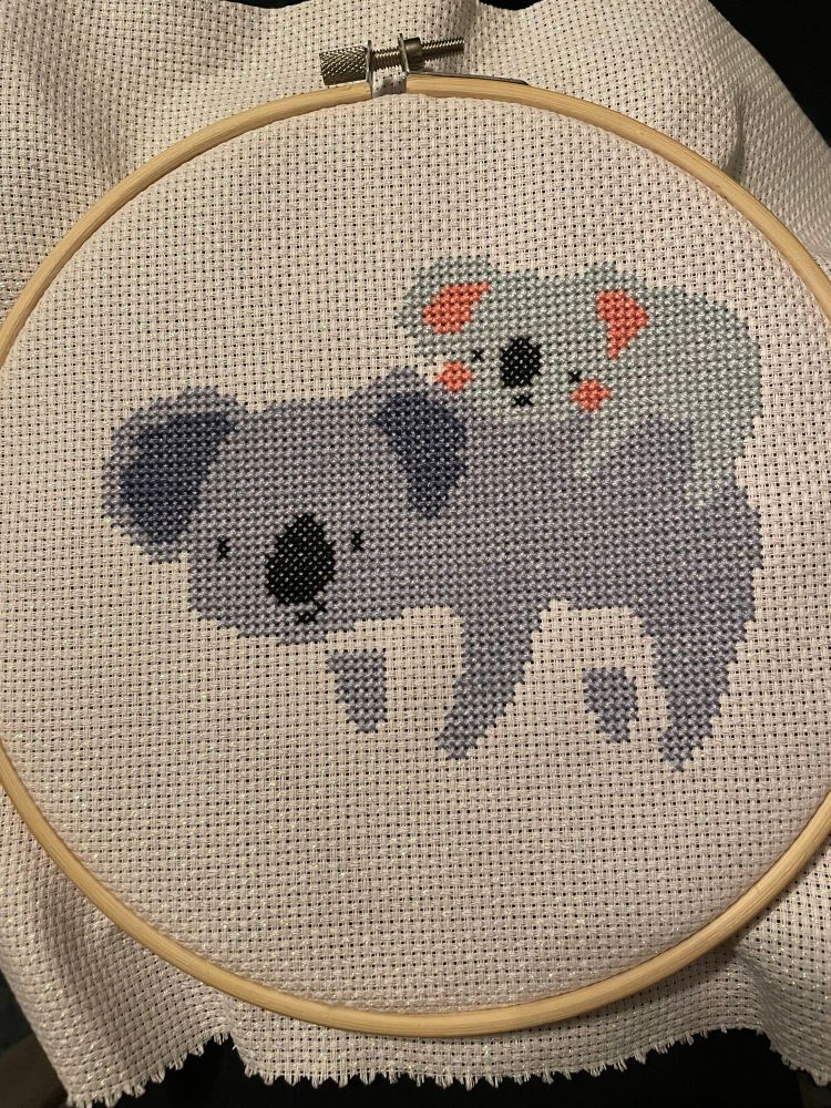 A light cross stitched blue baby koala rests on top of a darker blue mama koala on sparkly Aida fabric in a hoop. 