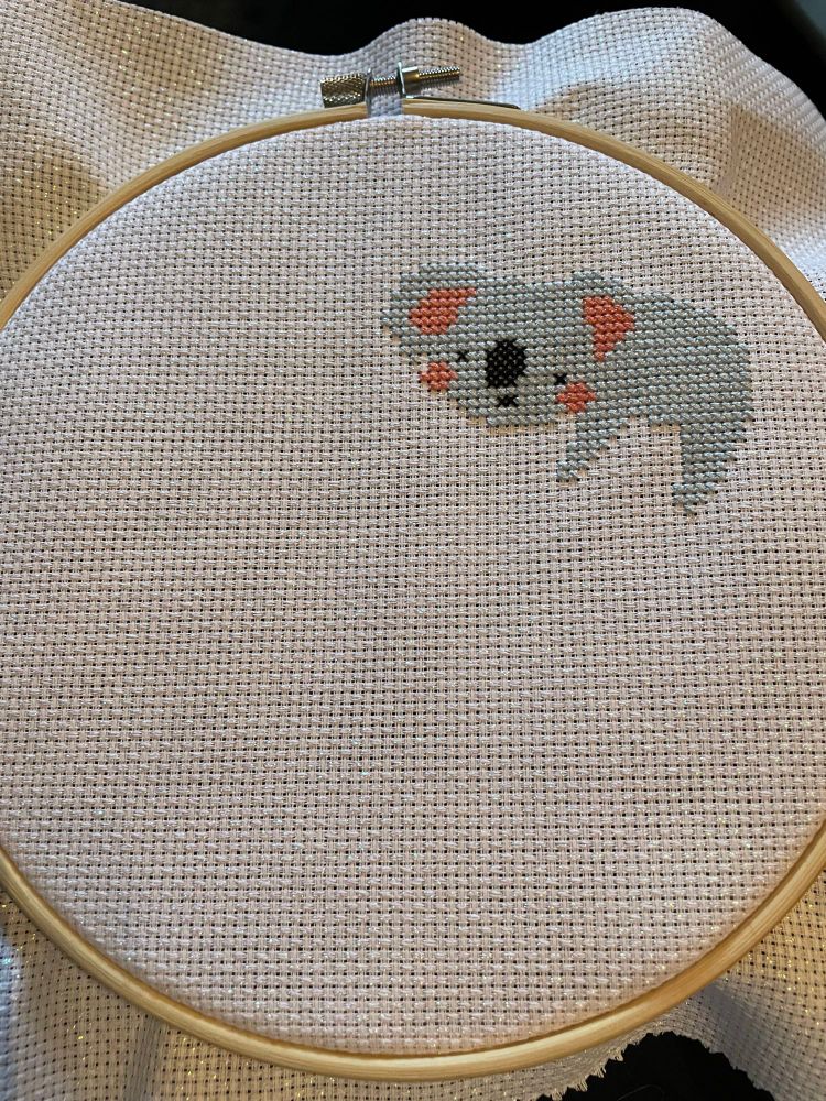 A cross stitched baby koala on a piece of Aida in a hoop. The baby koala is light blue with pinkish accents. 