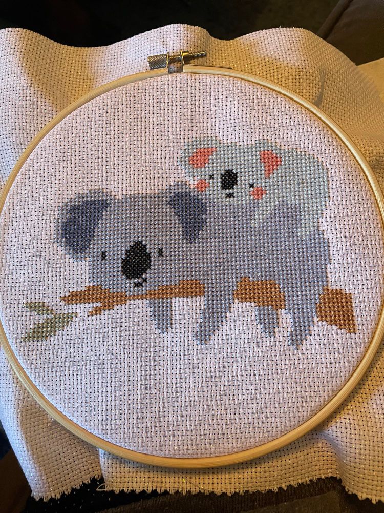 A baby koala laying on top of a mama koala who is laying on a branch with green leaves at the end. Cross stitch on sparkly white Aida in a hoop. 