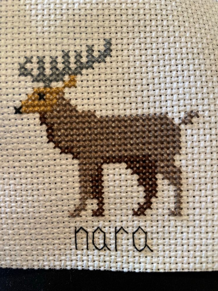 A cross stitch on white Aida of a brown deer with a tan face and gray antlers with the word “Nara” backstitched in black thread beneath it. 