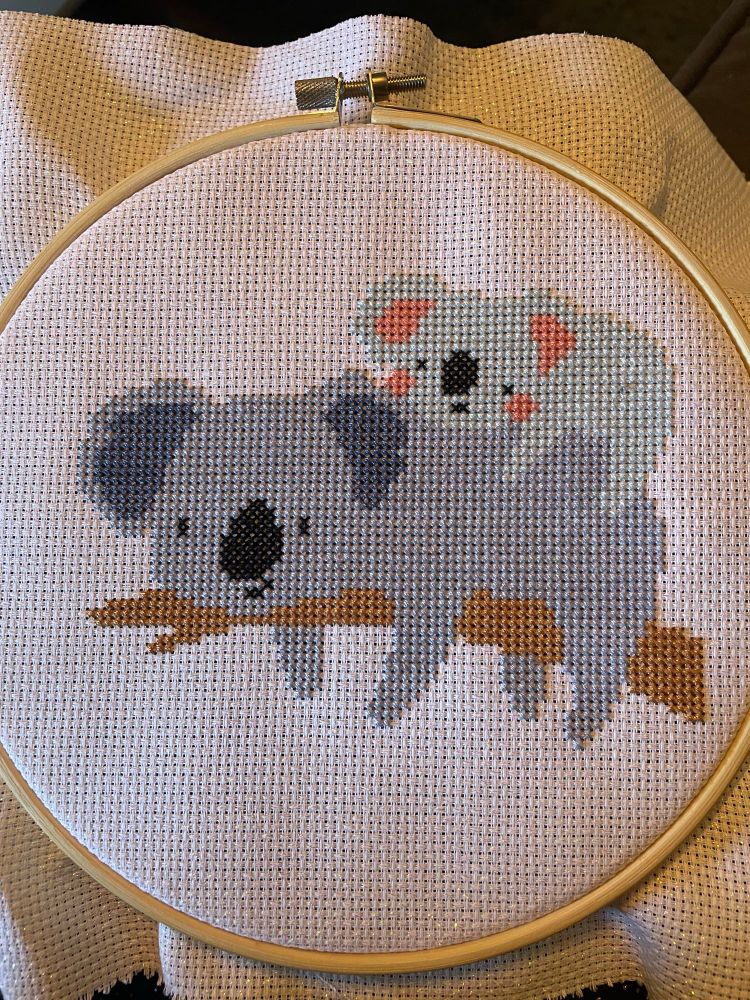 Baby koala resting on mama koala who is resting on a branch. Cross stitch on sparkly white Aida surrounded by a hoop. 