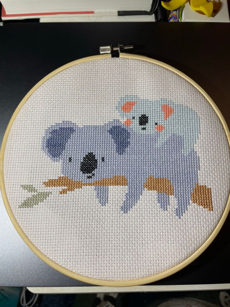 A cross stitch in a hoop on sparkly white Aida with two koalas on a branch- a dark blue patent and a light blue baby 