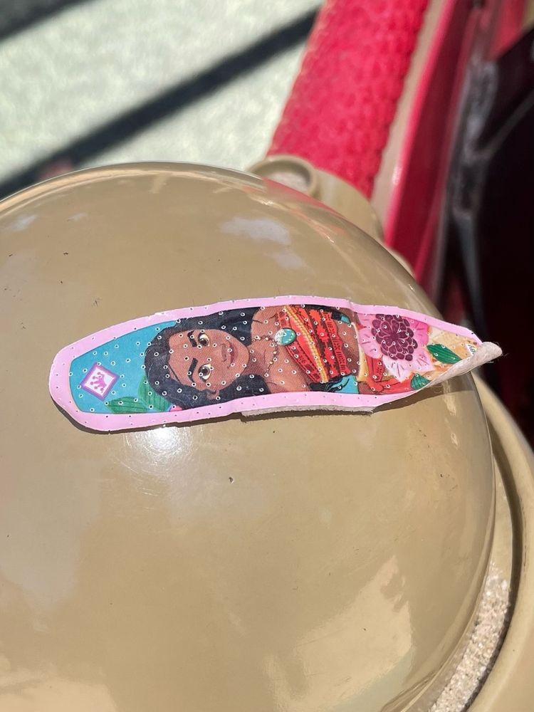 A used, pink-framed Band-Aid of the Disney character Moana, in a floral dress and red halter, is stuck onto a beige dome cover on a children’s playground set.