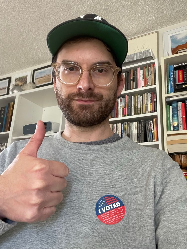 White guy in a grey sweatshirt with a red and blue I Voted sticker gives a big thumbs up for completing his civic duty. In the background are too many fucking Criterions.