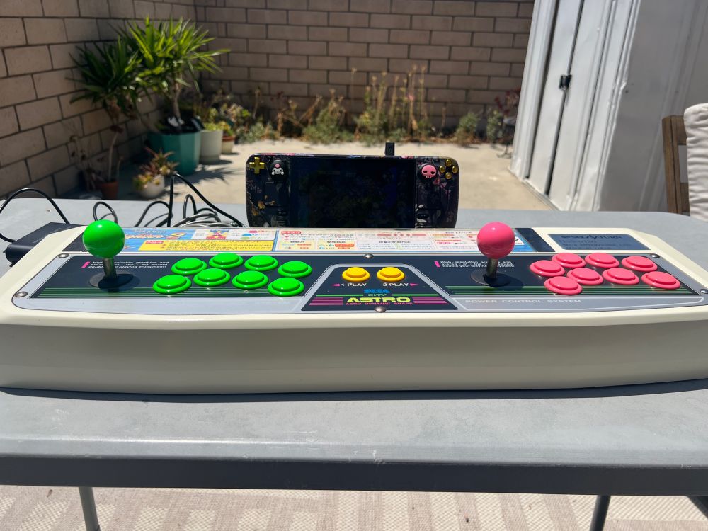 A Sega Saturn Virtua Stick Pro (a two-player arcade stick that recreates the Astro City cab's control deck) is connected to a customized Steam Deck. Both are placed on top of a folding table in a sunny back yard.