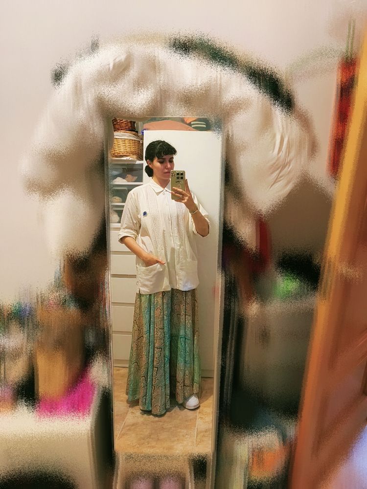 Selfie en el espejo para enseñar un conjunto de ropa que incluye: guayabera blanca con bolsillos bajos, un pantalón estilo falda de vuelo con estampados verdes y dorados, y unas deportivas blancas.