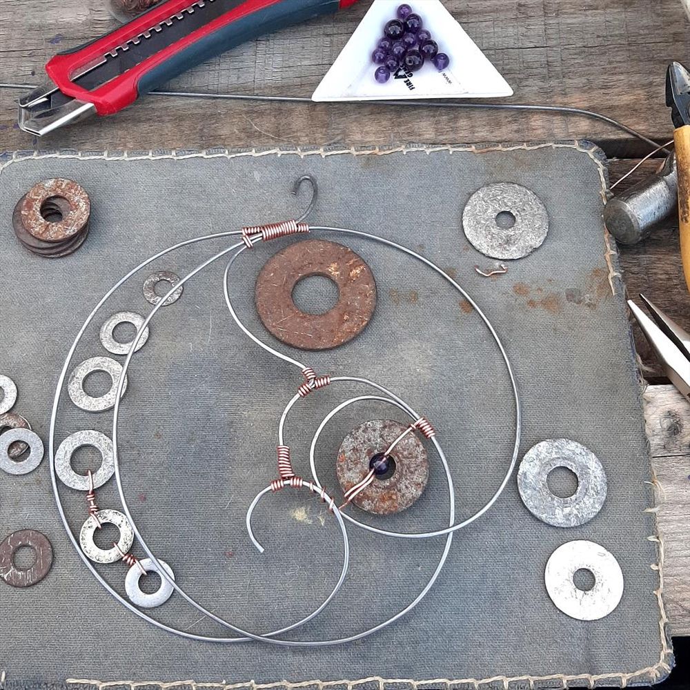 Three lengths of steel wire sit on my workbench. Each has been bent into a simple curlicue, and they've been placed so that together they form a pleasing shape, and joined together with copper wire wrapped around them.  A line of old beat-up washers runs up the left side; some of those have also been joined with copper wire. Other washers are scattered around, along with purple beads and a few jewelry tools.