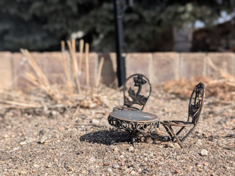 Macro photo of a miniature antique-style patio table and chairs, big enough to probably fit on an outstretched hand, placed in a barren garden bed 