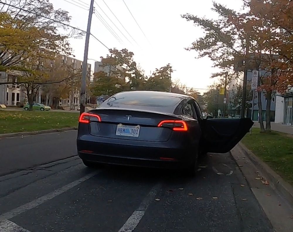 A grey car stopped in a bike lane in Halifax/Kjipuktuk, Nova Scotia. The car was identified as an Uber by the passenger who got out direcly into my path as I cycled on the bike lane. A no stopping and no parking sign can be seen to the right on the verge and a faded painted bicycle is just visible under the open car door