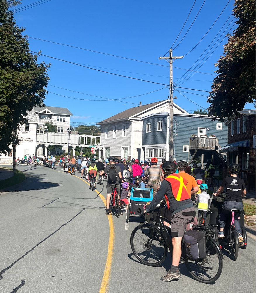 A long line of kids and their families begin to pass through an intersection in Dartmouth, Nova Scotia. The sun is hining and there is a blue sky at the top of the picture, buildings and trees.  It's a KidicalMassHFX bike ride, a joyful protest for safe streets for children to move around safely and independently.