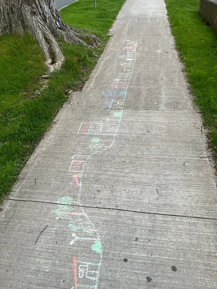 A photo of a sidewalk (aka pavement) on a city street with a child's chalk dawing of houses interspersed with trees along a squiggly line that presumably represents the street boundary. Conspicuously missing fom the drawing are all the parked cars that are stored for free and that prevent children from being able to roam independently around their neighbourhoods. 