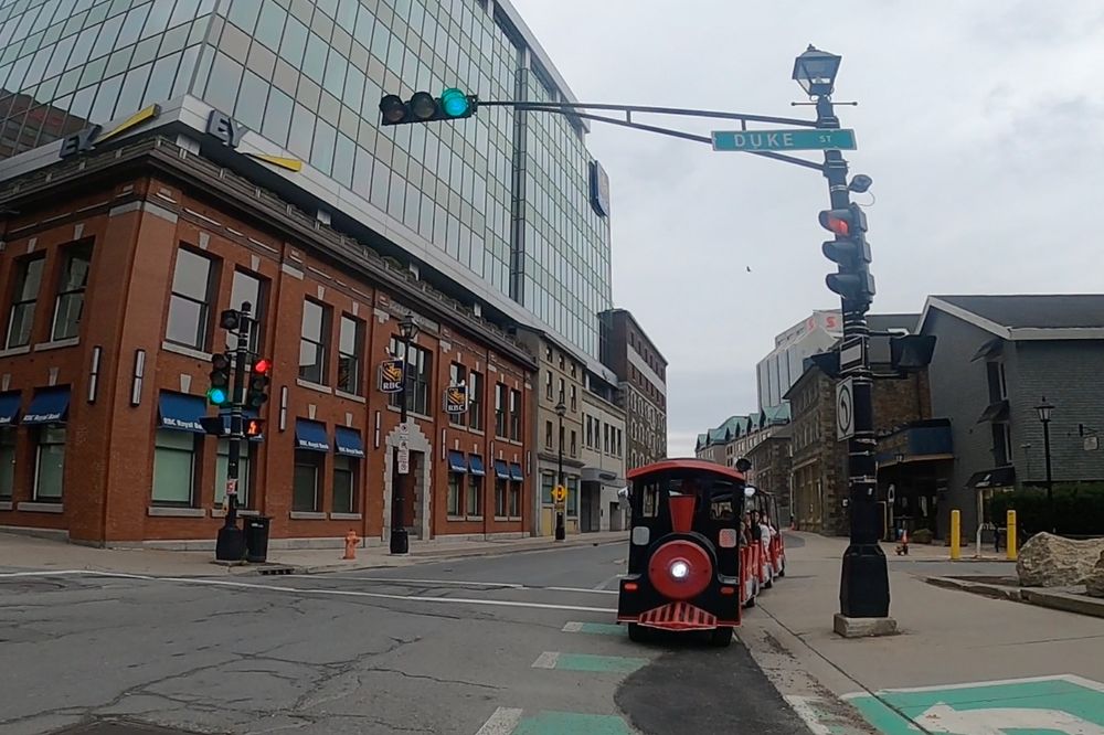 An image of a city street (downtown Halifax/Kjipuktuk) from the POV of a person riding in the bike lane. Coming the wrong way in the bike lane is a black and red model train with a driver and people sitting in the passenger cars behind it. The train is captured by the camera as the cyclist moves into the street to get past. There are buildings in the background.