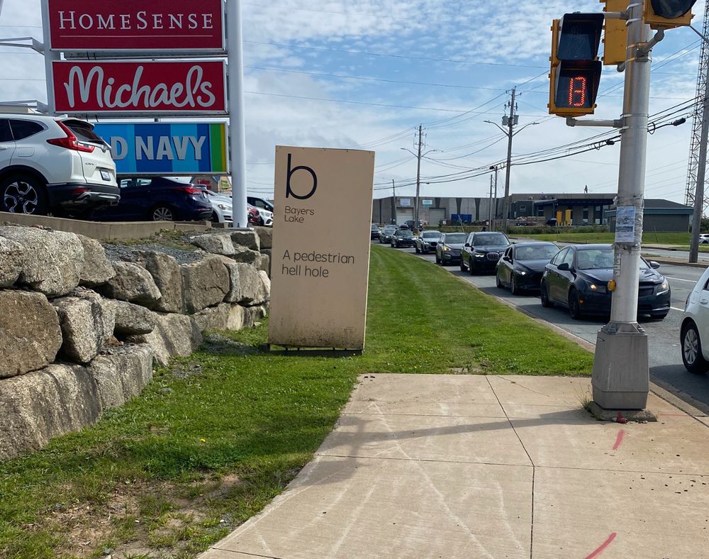 A photo taken in a out-of-town, car-centric shopping centre (Bayers Lake, Kjipuktuk/Halifax). It shows a cloudy blue sky, a line of cars at a traffic light, some shop signs and a sign that reads "Bayers Lake". Just in front of the sign, the pedestrian sidewalk ends but there is a faint path in the grassy verge that suggests that people walk along the verge despite the lack of infrastructure. Added to the sign are the words "A pedestrian hell hole" in recognition of the car-centric design that means anyone walking, cycling or using transit is literally pushed to the margins or excluded altogether. 