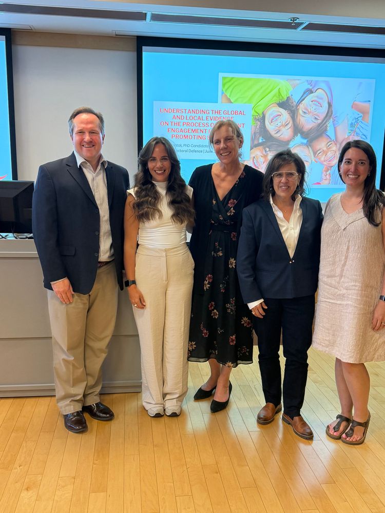 Image of a presentation on screen in a classroom. There are 5 people standing in front of the presentation. They are all smiling at the camera. The occasion is a PhD defence with the candidate and committee shown in the photo.