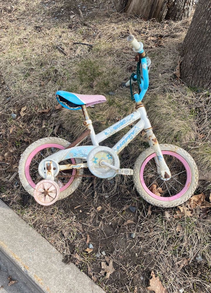 A children's bike lies on a verge on the side of a street. The bike is white, pink and blue with training wheels. The grass verge is muddy & leaf strewn in the early spring & there are trunk of trees visible in the background