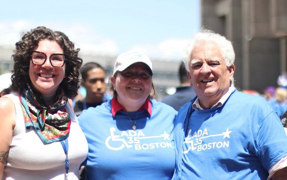Three smiling people posing together at what appears to be an outdoor community event. Two people are wearing blue “ADA 35 BOSTON” t-shirts, while the person on the left wears a colorful floral scarf. The setting appears to be in an urban area with buildings visible in the background.