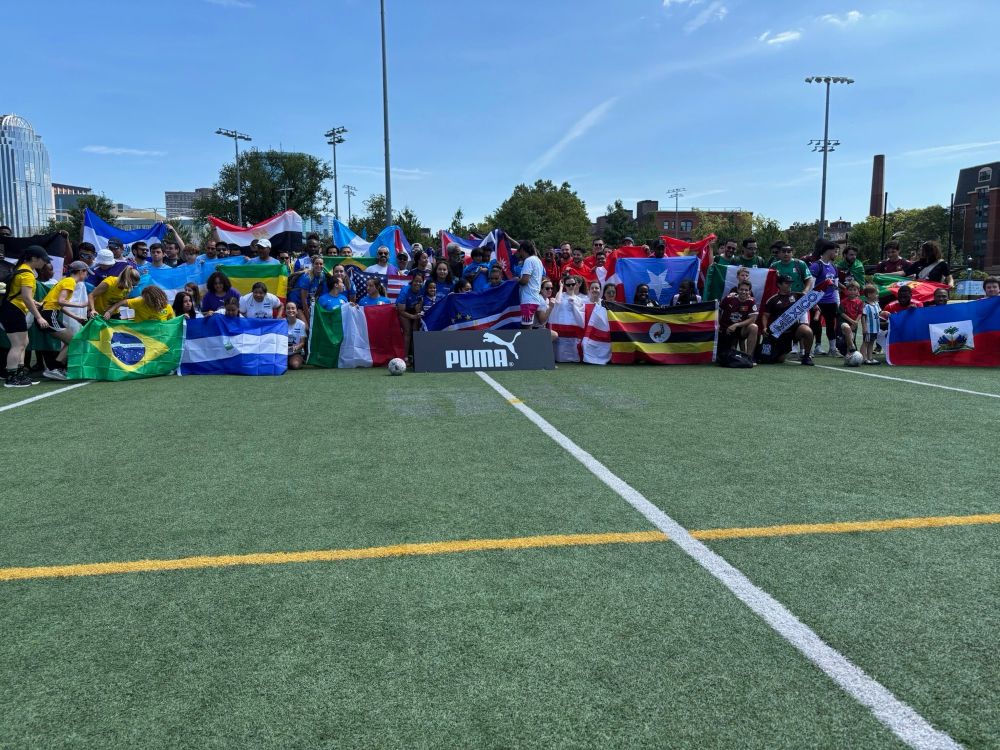 A diverse group of people poses with flags from various countries including Brazil, France, Italy, Puerto Rico, Haiti, and others on a soccer field. The colorful display of international flags creates a vibrant backdrop along the sideline, with participants gathered for what appears to be a multicultural soccer event.