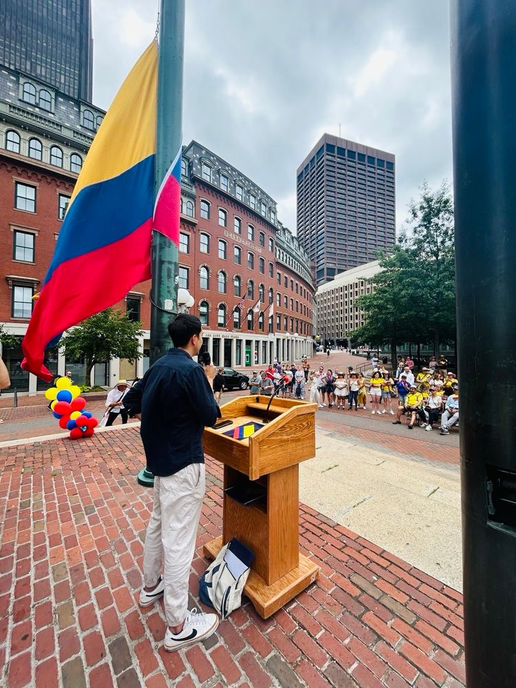A person in dark clothing stands at a wooden podium speaking to a gathered crowd at Boston City Hall Plaza. Behind them, a large Colombian flag with yellow, blue, and red horizontal stripes is prominently displayed on a flagpole. Colorful balloons and the plaza’s characteristic brick surface are visible, with historic and modern buildings forming the backdrop.