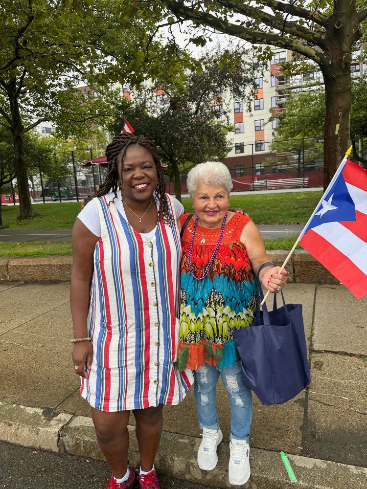 Ruthzee Louijeune in her striped dress poses with an older woman wearing a colorful patterned top and jeans. The older woman is holding a Puerto Rican flag. They’re standing on a sidewalk with apartment buildings and green space visible in the background.