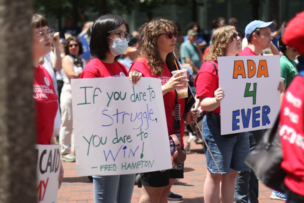 A group of people at an outdoor rally holding signs. One person wearing a face mask and red shirt holds a sign reading “If you dare to struggle, you dare to win! - Fred Hampton.” Another person holds a sign that says “ADA 4 EVER.” The crowd includes people wearing various colored shirts, and the scene appears to be on a brick-paved area with trees in the background.