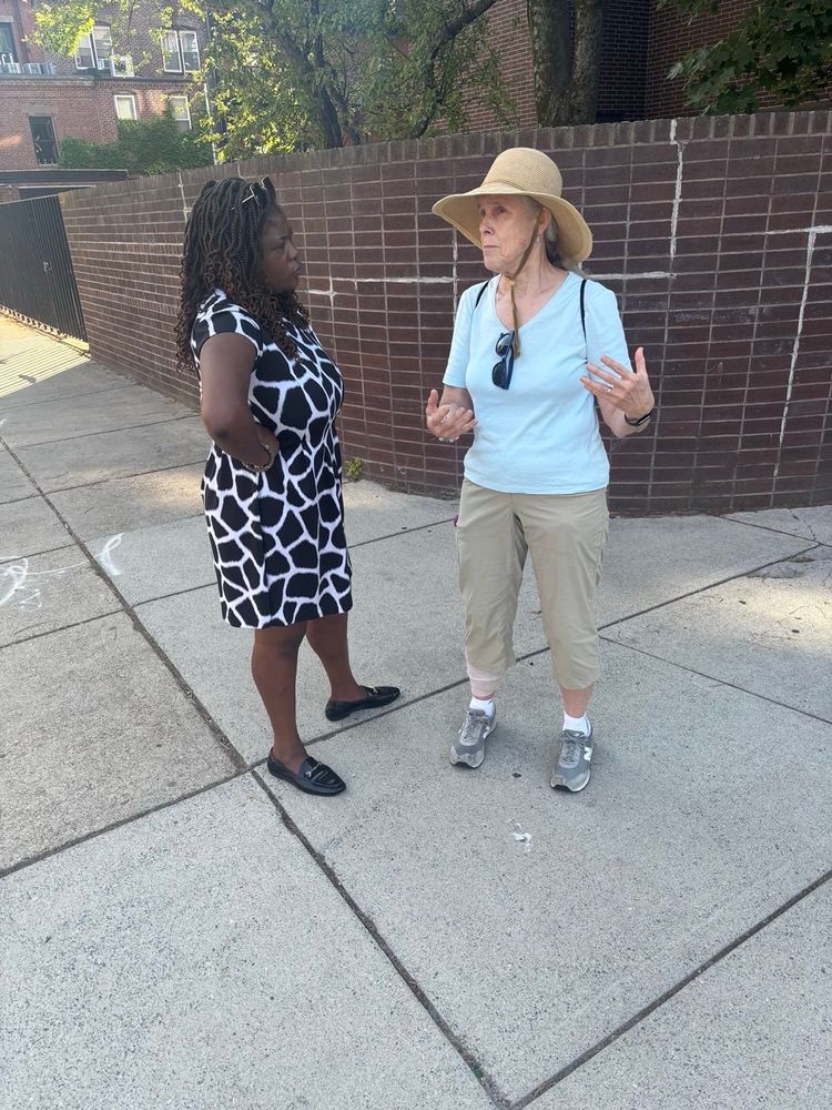 Ruthzee Louijeune and a woman having a conversation on a sidewalk. One woman wears a black and white geometric patterned dress, while the other wears a light blue top, beige pants, and a wide-brimmed hat. They appear to be engaged in discussion near a brick wall.