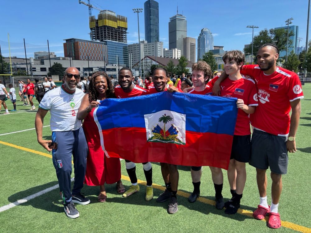 A group holds up the Haitian flag prominently in front of a city skyline featuring tall buildings and construction cranes. The group includes people wearing various soccer jerseys and casual clothing, gathered on the turf field for a photo opportunity celebrating Haitian heritage.